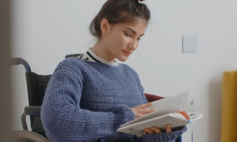 mlada devojka sa invaliditetom sedi u kolicima i lista knjigu